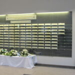 Lasivitriinissä useita vaakasuoria, suorakulmaisia muistolaattoja. Vitriinin yläpuolella on kaksi valaistu kylttiä. Vitriinin edessä on valkoisella liinalla peitetty pöytä, jonka päällä on kukka-asetelmia. Pöydän vieressä oikealla on tummansininen lippu.