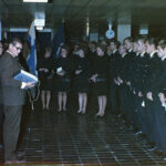 Värillinen kuva tilaisuudesta, jossa rivi tummiin univormuihin pukeutuneita ihmisiä seisoo. Edessä siviilipukuinen mies lukee papereita Suomen lipun vieressä.