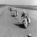 Mustavalkoinen kuva harjoituksesta, jossa joukko moottoripyöräpoliiseja ajaa jonossa asfalttitiellä kartioiden lomassa. Poliiseilla on kypärät ja moottoripyörissä sivulaukut.