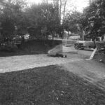 Mustavalkoinen kuva puistosta, jossa hiekkatie haarautuu. Tiellä henkilö makaa vatsallaan. Etualalla on nurmikkoa, ja taustalla näkyy auto ja puita.