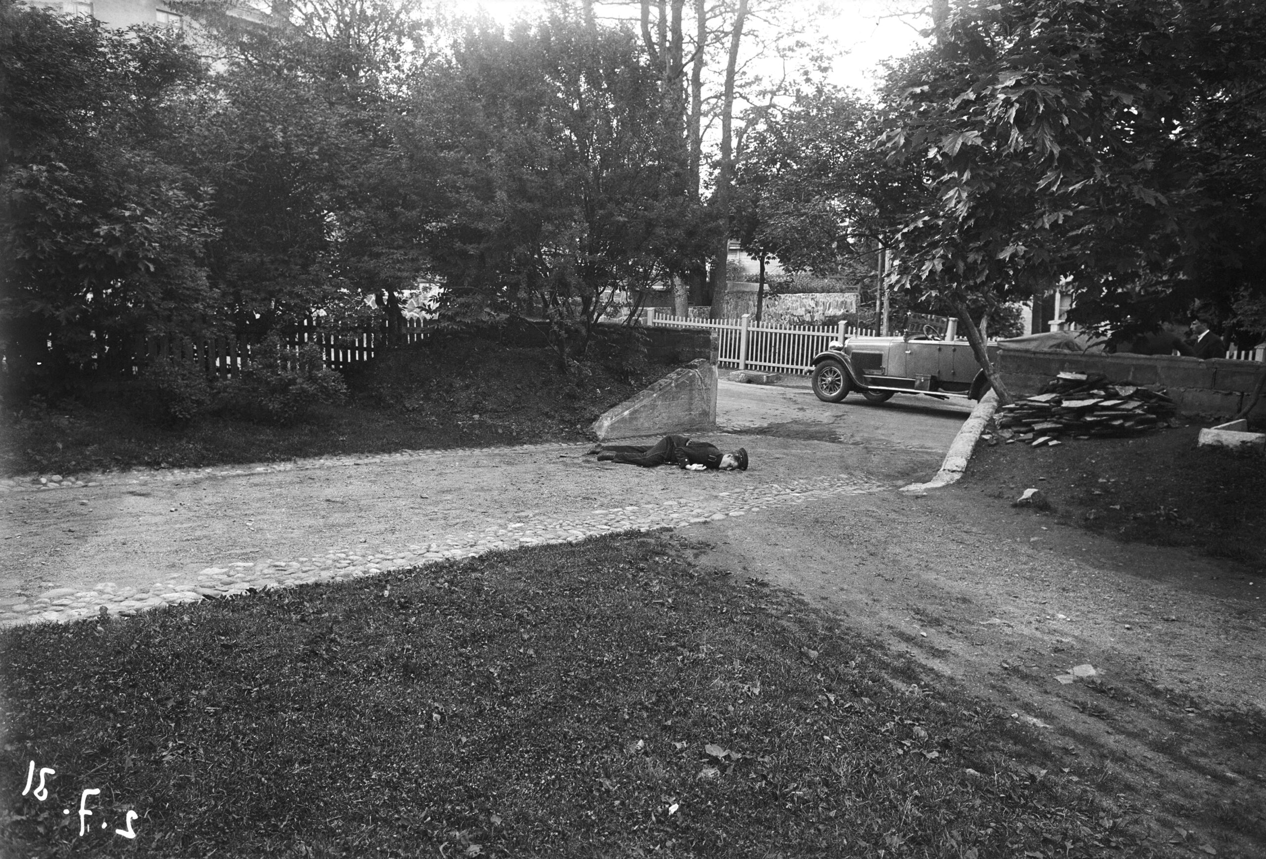Mustavalkoinen kuva puistosta, jossa hiekkatie haarautuu. Tiellä henkilö makaa vatsallaan. Etualalla on nurmikkoa, ja taustalla näkyy auto ja puita.