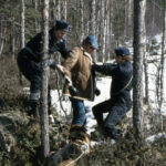 Kaksi virkapukuista poliisia pitää kiinni miehestä metsässä, jossa on hieman lunta. Etualalla maassa seisoo saksanpaimenkoira.