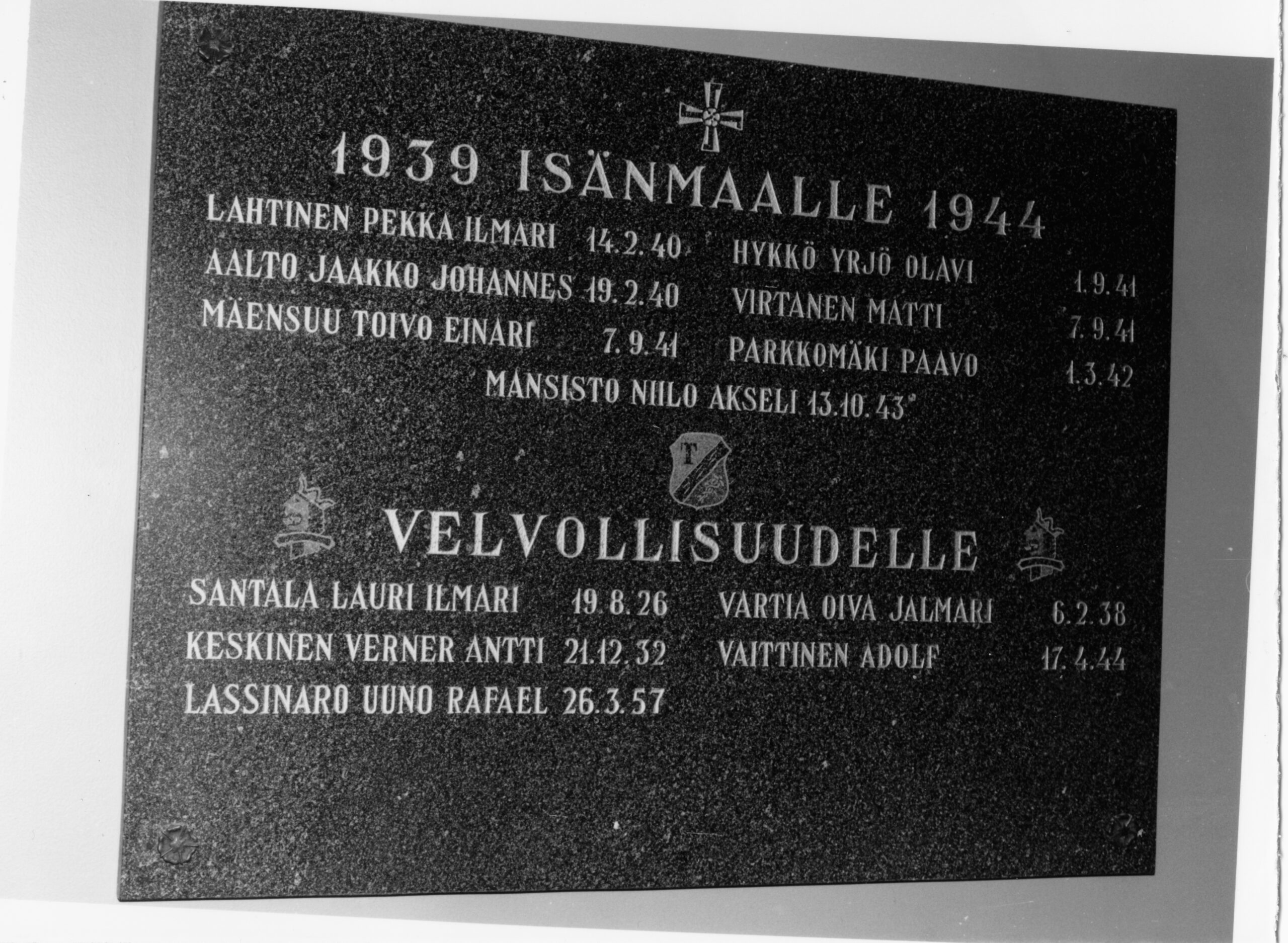 Mustavalkoinen lähikuva tummasta muistolaatasta, jossa on kaiverrettu risti ja tekstiä. Laatta on jaettu kahteen osaan, joiden otsikot ovat "1939 ISÄNMAALLE 1944" ja "VELVOLLISUUDELLE". Otsikoiden alla on lueteltu nimiä syntymäaikoineen ja kuolinpäivineen.