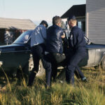 Värillinen kuva, jossa kaksi virkapukuista poliisia pitelee kolmatta henkilöä mustan auton vieressä. Taustalla näkyy rakennuksia.