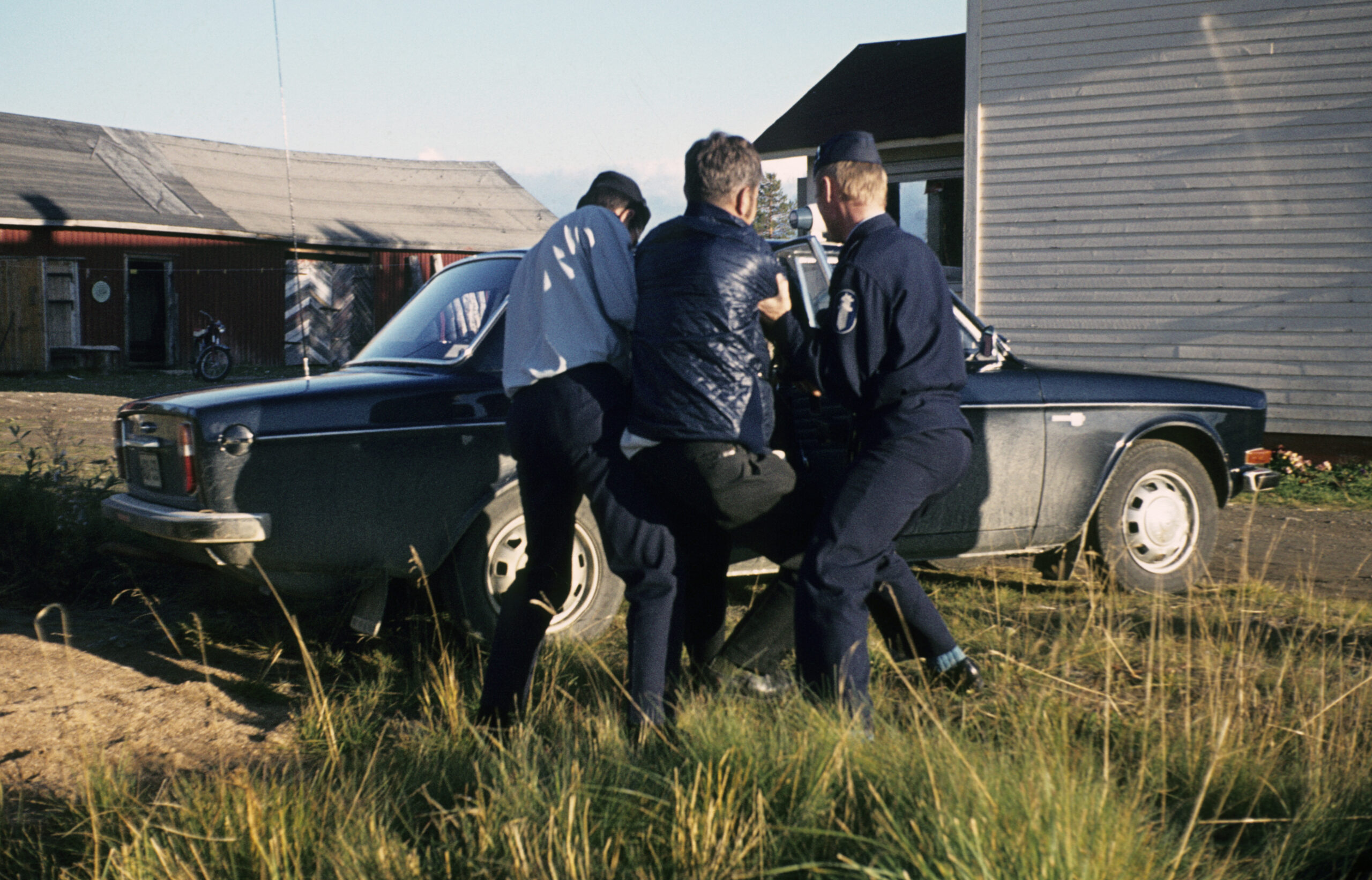 Värillinen kuva, jossa kaksi virkapukuista poliisia pitelee kolmatta henkilöä mustan auton vieressä. Taustalla näkyy rakennuksia.