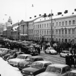 Mustavalkoinen kuva hautajaissaattueesta, jossa etummaisena auto ja auton ympärillä virkapukuisia poliiseja. Auton takana virkapukuisia poliiseja pitkässä jonossa. Osa kantaa soittimia. Kuva on talvinen. Taustalla näkyy suuria, klassisia rakennuksia, joiden katoilla on lunta.