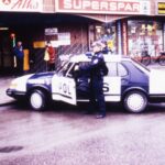 Värillinen kuva, jossa poliisi seisoo kadulle pysäköidyn poliisiauton vieressä. Kuljettajan ovi on auki. Taustalla näkyy kaupan sisäänkäynti.