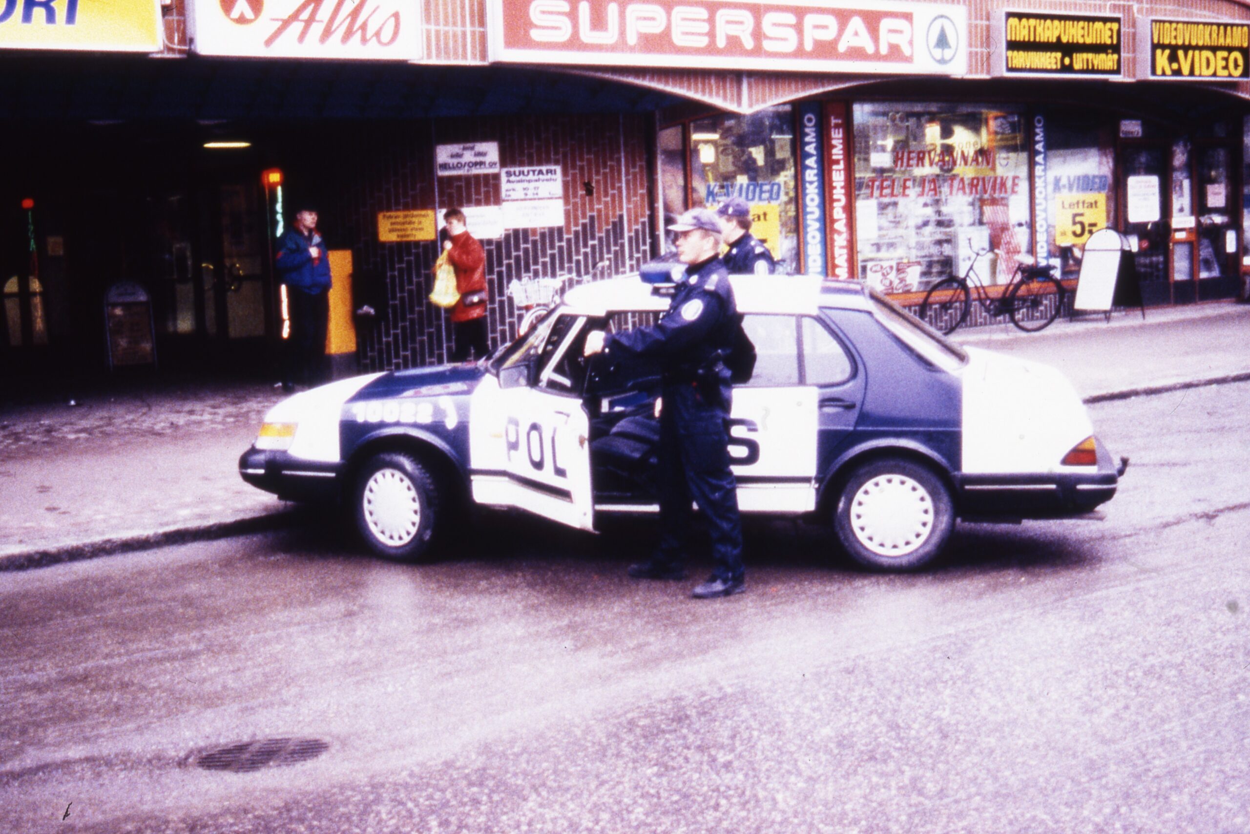 Värillinen kuva, jossa poliisi seisoo kadulle pysäköidyn poliisiauton vieressä. Kuljettajan ovi on auki. Taustalla näkyy kaupan sisäänkäynti.