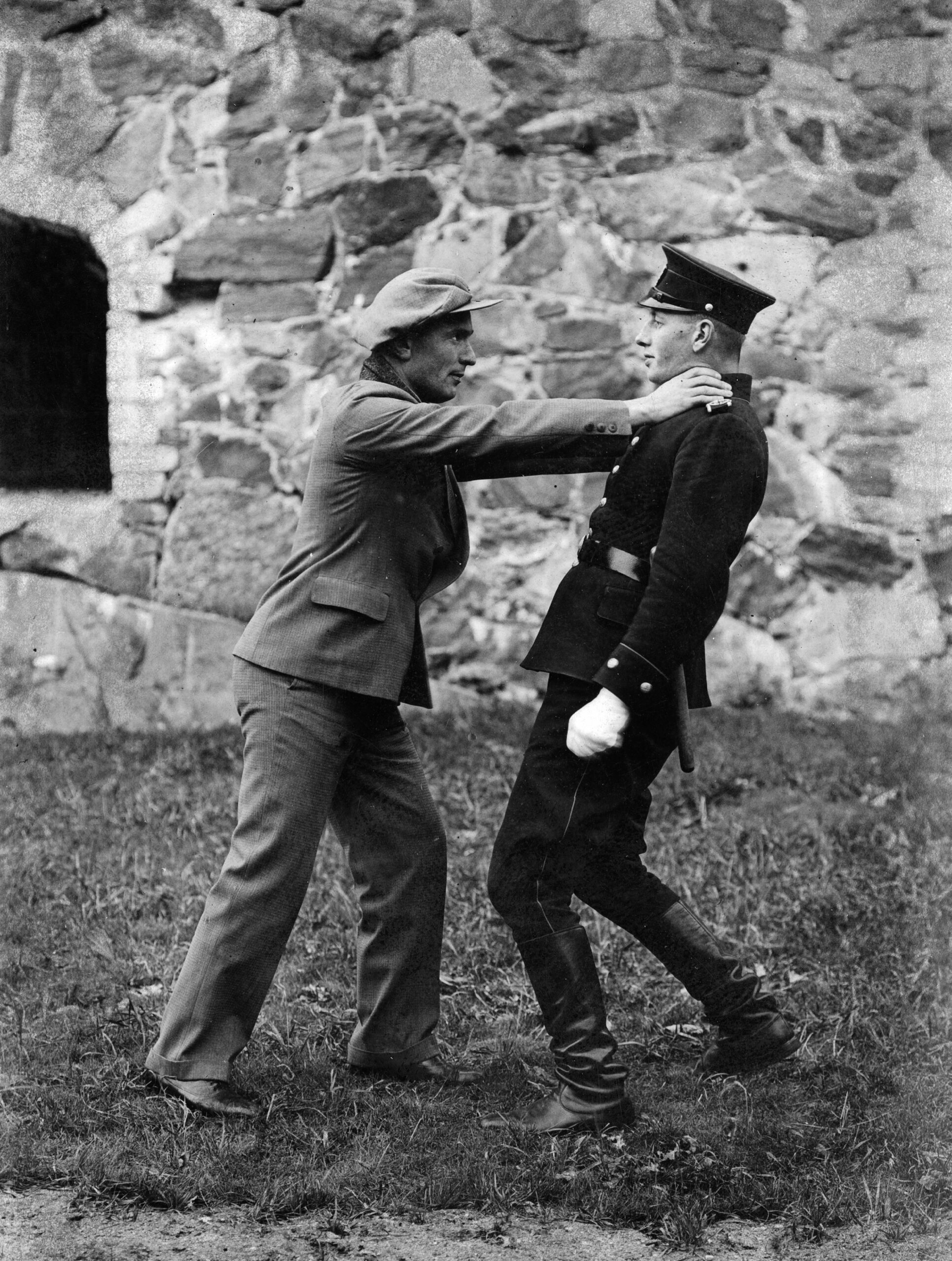 Mustavalkoinen kuva, jossa siviiliasuinen henkilö tarttuu käsillään virkapukuisen poliisin hartioihin. Taustalla näkyy kiviseinä.