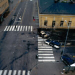 Ylhäältä otettu kuva kaupungin risteyksestä, jossa on suojateitä, yksi pahoin vahingoittunut vaalea henkilöauto ja kaksi poliisiautoa. Oikeapuoleisessa rakennuksia on myös vaurioita.