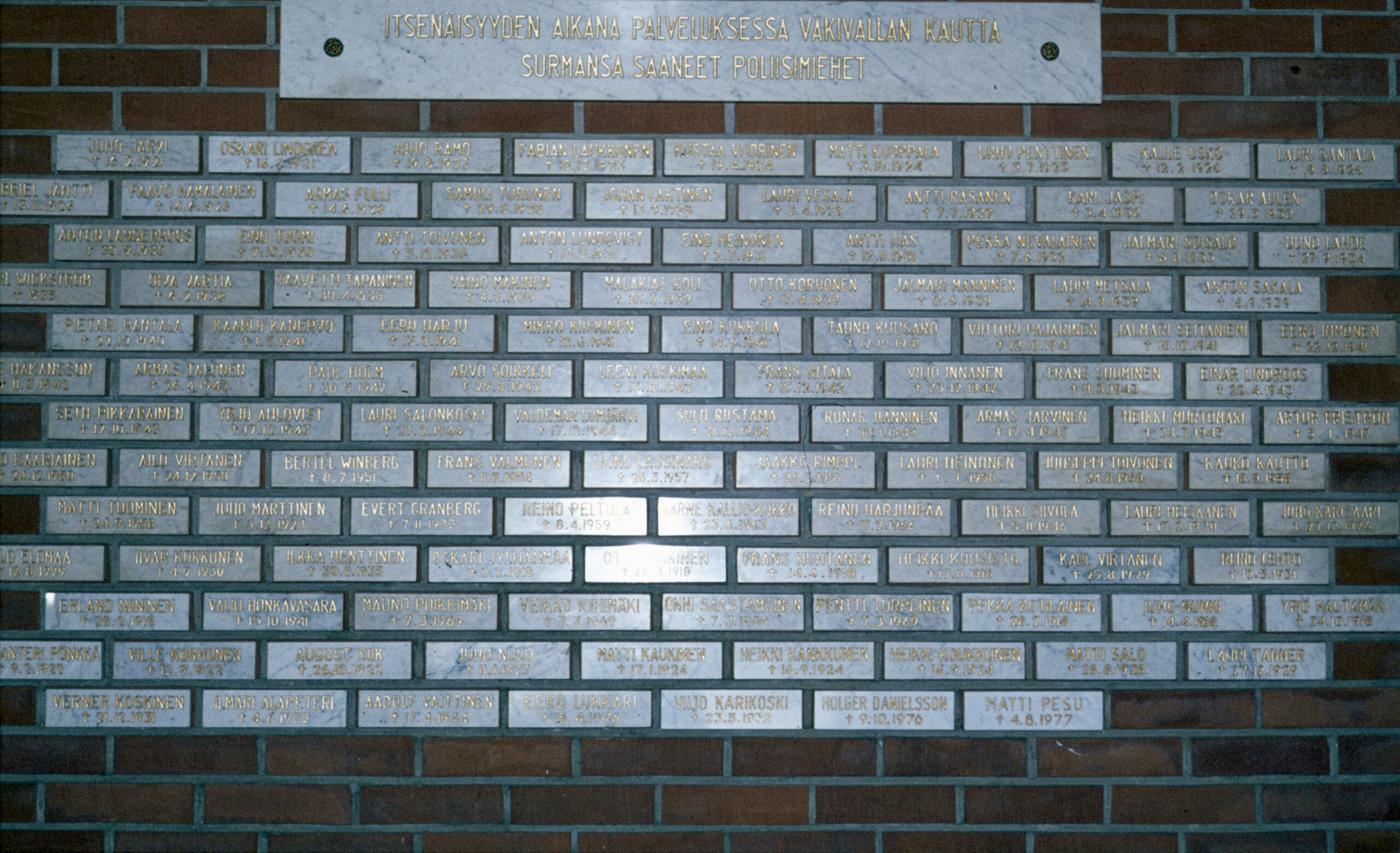 Tiiliseinään on kiinnitetty vaaleita laattoja, joissa on henkilön nimi ja kuolinaika. Päällä iso laatta, jossa teksti: Itsenäisyyden aikana palveluksessa väkivallan kautta surmansa saaneet poliisimiehet.