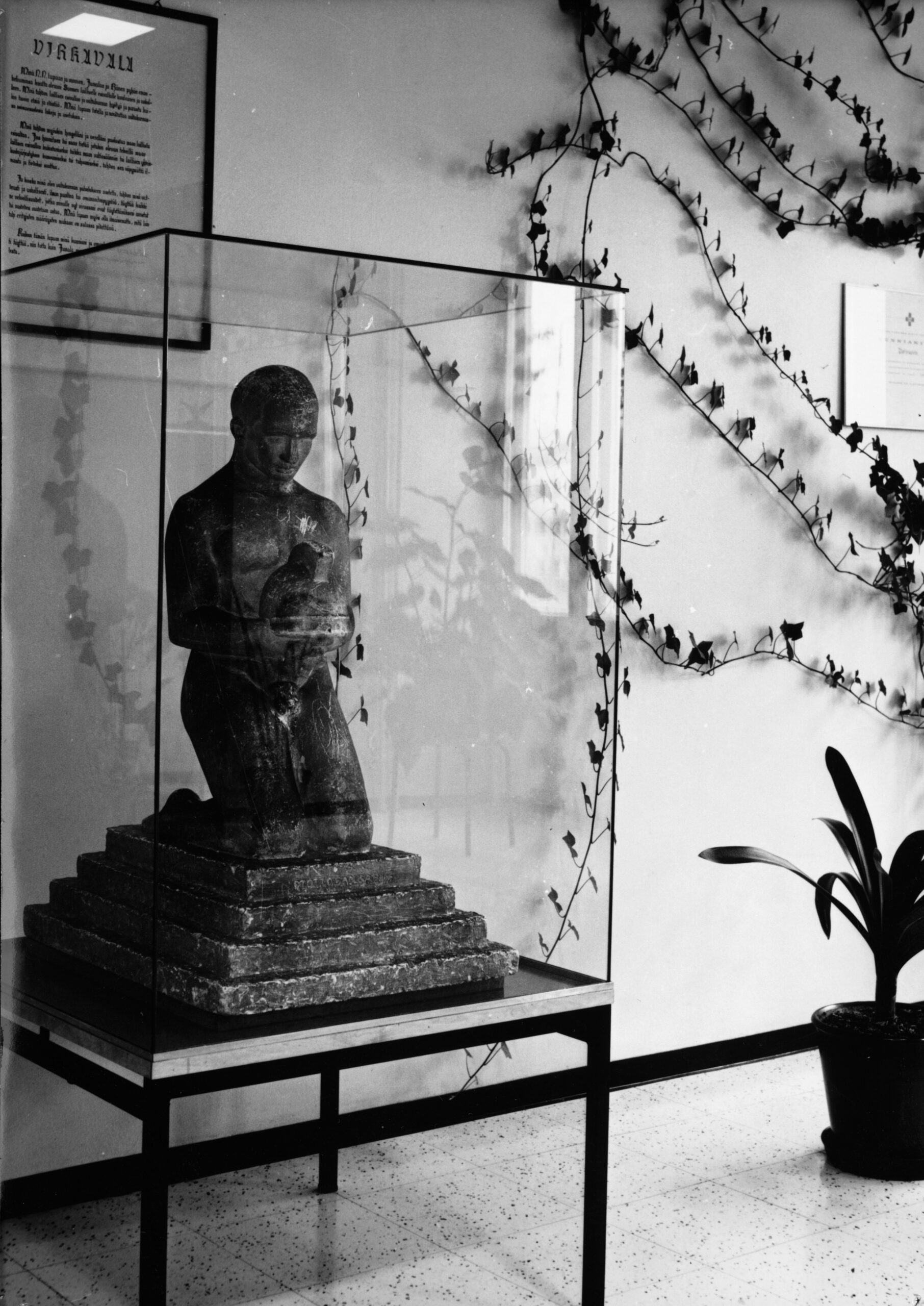 Kuvun sisällä veistos, jossa miesfiguuri on polvillaan portaiden päällä. Kädessä kypärä. Seinällä taulu, jossa tekstinä virkavala. Seinällä on köynnöskasvi.