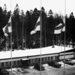 Mustavalkoinen talvikuva, jossa useita lippuja on laskettu puolitankoon rakennuksen edessä. Rakennuksen edessä on pysäköitynä useita autoja ja taustalla näkyy metsää.