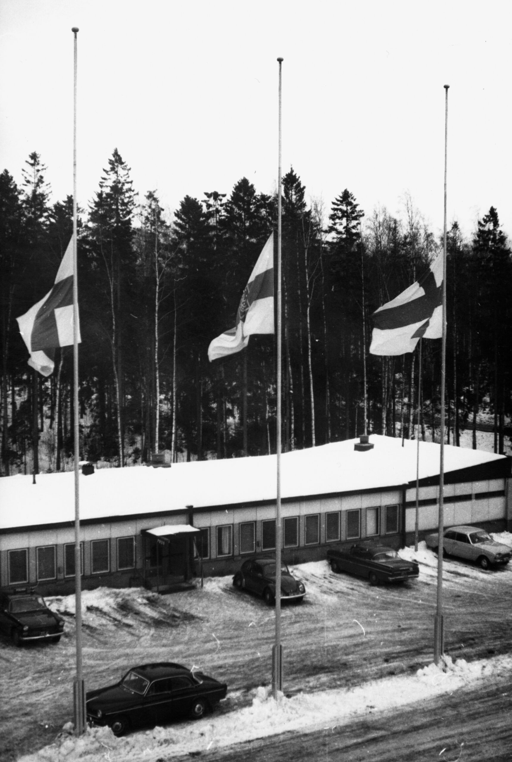 Mustavalkoinen talvikuva, jossa useita lippuja on laskettu puolitankoon rakennuksen edessä. Rakennuksen edessä on pysäköitynä useita autoja ja taustalla näkyy metsää.