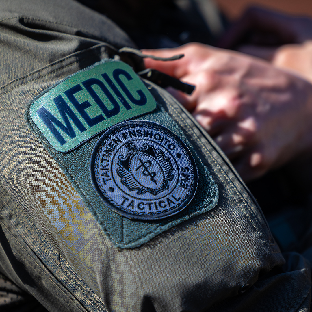 Lähikuva hihamerkistä, jossa lukee "MEDIC" ja toinen pyöreä tunnus, kiinnitettynä harmaanvihreään kankaaseen.