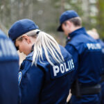 Lähikuva kahdesta virkapukuisesta poliisista selkäpuolelta. Etualalla olevan naisen tummansinisen takin selässä lukee valkoisilla kirjaimilla "POLIISI". Toinen poliisi näkyy taustalla epätarkkana.