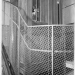Mustavalkoinen kuva, jossa portaita kiertää metalliverkosta tehty kaide. Taustalla näkyy hissin seinä ja ovi, jonka ikkunassa on reikä.