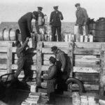 Mustavalkoinen kuva, jossa ryhmä miehiä on laivan kannella ja laiturilla. He siirtävät tynnyreitä.