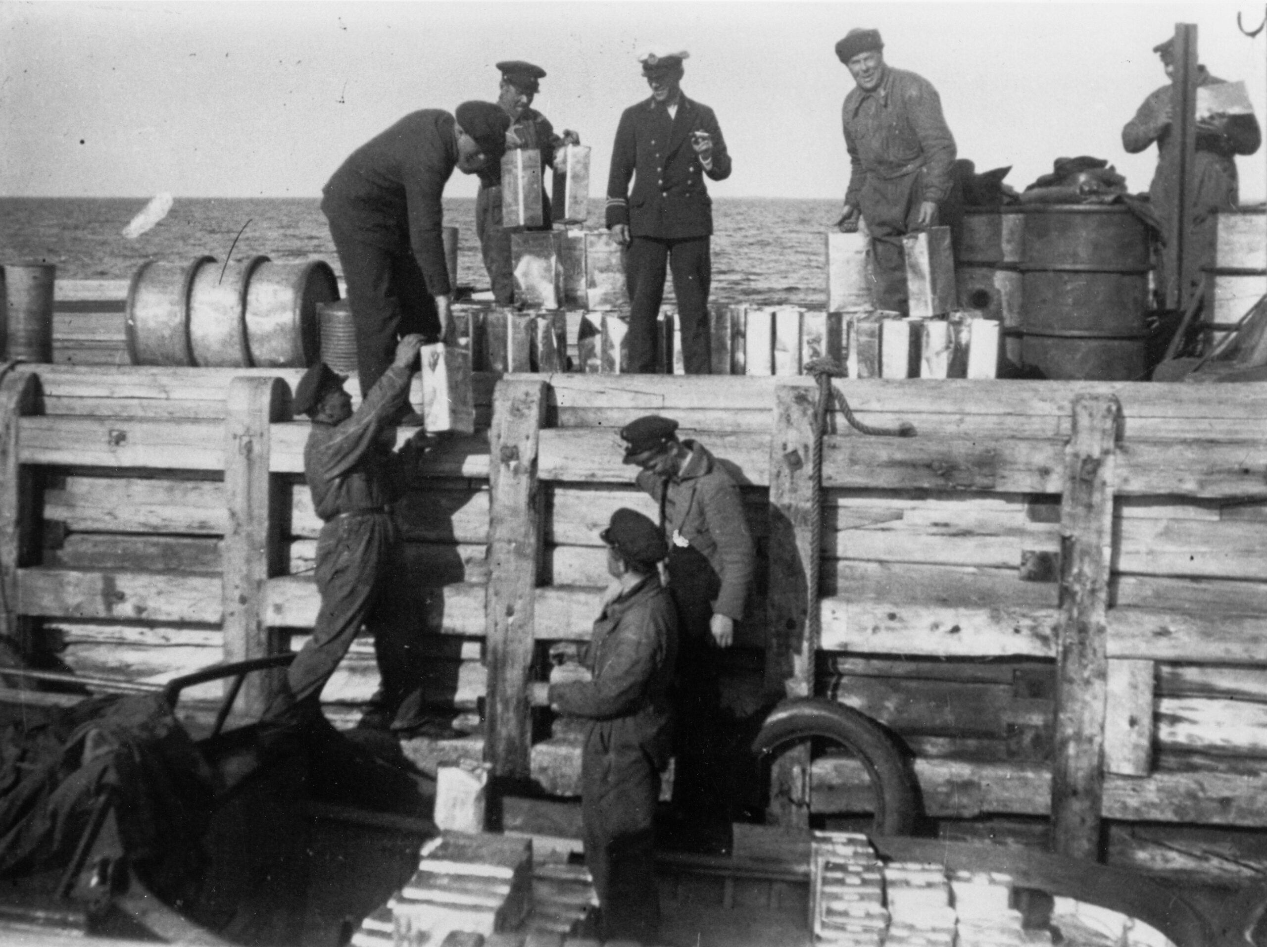 Mustavalkoinen kuva, jossa ryhmä miehiä on laivan kannella ja laiturilla. He siirtävät tynnyreitä.